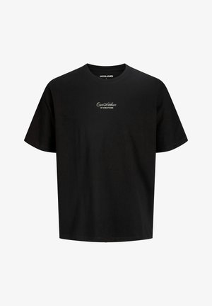 Garçon souriant debout, portant un t-shirt noir à manches courtes, un pantalon beige à jambes larges et des baskets blanches sur un fond blanc uni.