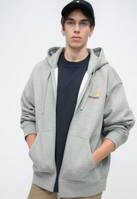 Siva dukserica na zip s opuštenim krojem, velikim prednjim džepovima i rebrastim manžetama. Na lijevoj strani prsnog dijela ima mali izvezeni Carhartt logo.