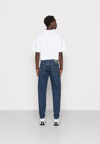 T-shirt bianco a maniche corte abbinato a jeans in denim blu scuro; i jeans presentano un taglio classico, tasche posteriori e un'etichetta del marchio sulla cintura.