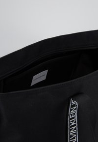 Czarna torba tote z logo Calvin Klein Jeans na uchwycie i etykietą wewnątrz, częściowo otwarta, pokazująca zapinaną kieszeń.