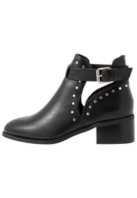 Bota de tobillo de cuero negra con tachuelas plateadas, corte lateral, correa ajustable con hebilla y tacón bajo de bloque.