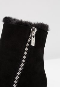 Bottine en daim noir avec une fermeture éclair argentée dotée d'une tirette de marque et un revers doux et semblable à de la fourrure le long du bord supérieur.