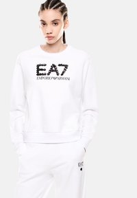 Sudadera blanca con cuello redondo, que presenta un logo texturizado en negro "EA7" y el texto "EMPORIO ARMANI" en una tipografía elegante. Tejido suave, corte relajado.