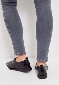 Chaussures noires en cuir avec une texture lisse et des bouts arrondis, présentant un détail de couture subtil et un talon bas et plat. Associées à un jean slim gris.