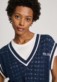 Tamsiai mėlynas megztinis su tekstūruotu raštu ir balta apdaila. Su V formos iškirpte ir "PEPE JEANS" siuvinėta ant krūtinės. Dėvimas ant baltos marškinės.