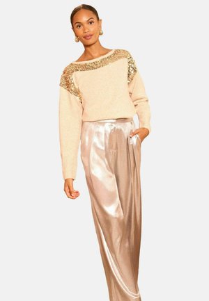Frau in einem beigen Pullover mit goldenen Pailletten an den Schultern und schimmernden, weiten Satin-Hosen, die mit einer Hand in der Tasche steht.