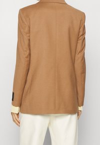 Brauner maßgeschneiderter Blazer mit glatter Textur, einfacher Belüftung und scharfen Revers. Weiße Hose ist am unteren Ende sichtbar, mit strukturierten Bündchen, die herausblitzen.