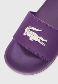 Violet spalvos slaidinės šlepetės su tekstūruota dirželiu, ant kurio yra baltas krokodilo logotipas. Lygus padas ir apvalus padas suteikia patogumo.