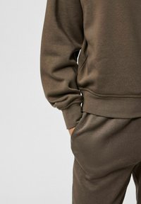 Set di pantaloni e felpa in verde oliva realizzati in tessuto morbido e liscio, con polsini e vita a coste. Presenta tasche laterali.