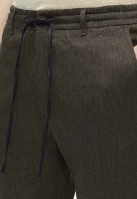 Pantalon en lin olive foncé avec une taille à cordon noir et des poches latérales, porté avec un haut beige rentré.