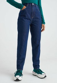 Džíny s vysokým pasem modré barvy s rovnými nohavicemi, s bočními kapsami a zapínáním na předním díle. Nosené s zeleno-bílými teniskami.