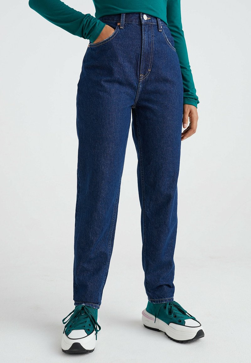 Džíny s vysokým pasem modré barvy s rovnými nohavicemi, s bočními kapsami a zapínáním na předním díle. Nosené s zeleno-bílými teniskami.