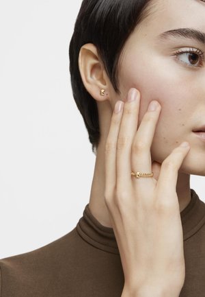 Goldfarbene Steckerohrringe und ein passender Ring mit strukturiertem Design, getragen von einer Person mit kurzen Haaren und einem braunen Oberteil.