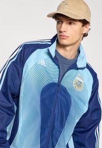 Μπλε και τιρκουάζ ζακέτα με κάθετες ρίγες, με κεντημένο λογότυπο και τρεις λευκές ρίγες στα μανίκια.