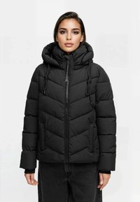 Mujer vestida con una chaqueta de plumas acolchada negra con un cuello alto, capucha y cordones, combinada con pantalones oscuros, contra un fondo liso.