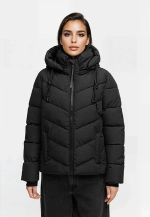 Woman wearing a black quilted puffer jacket with a high collar, hood, and drawstrings, paired with dark pants, against a plain background.