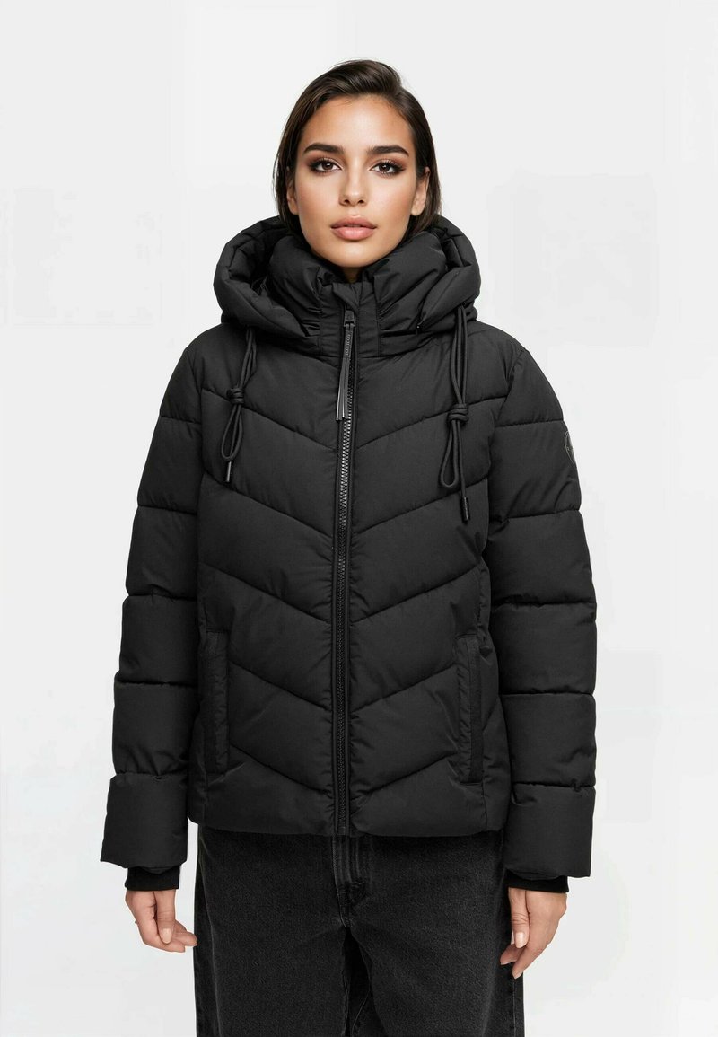 Mujer vestida con una chaqueta de plumas acolchada negra con un cuello alto, capucha y cordones, combinada con pantalones oscuros, contra un fondo liso.