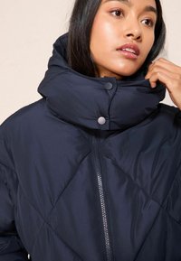 Námořnická puffer bunda s vysokým límcem, s diamantovým prošíváním, předním zipem a patenty na zavírání. Hladká textura.