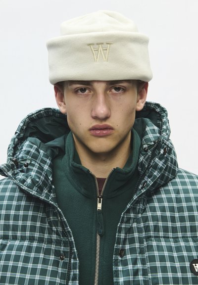 Jeune homme portant un bonnet en laine crème avec un logo brodé et une veste matelassée à carreaux verts sur une polaire verte zippée, regardant droit devant.