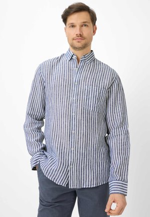 Mann trägt ein blau-weiß gestreiftes Hemd mit langen Ärmeln und dunkle Hose, steht mit einer Hand in der Tasche.