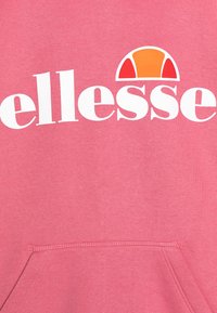 Růžová mikina s velkým bílým logem s nápisem "ellesse." Nad logem se nachází kulatý oranžový a červený grafický prvek. Měkká textura.