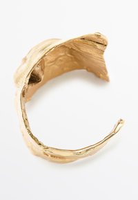 Bracciale rigido in oro con un design organico e testurizzato. Presenta un bordo ondulato e una finitura lucida, che evidenzia la sua superficie liscia e la forma irregolare.