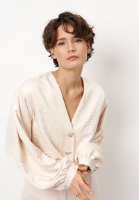 Lichtbeige blouse van textuurstof, diepe V-hals en grote knopen. Heeft puffed mouwen met geplooide manchetten.
