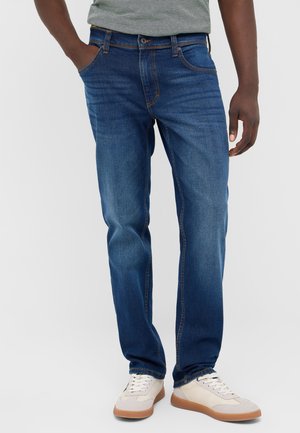 Mann mit blauen Jeans und weißen Sneakers mit Gummisohlen, der mit einer Hand in der Tasche steht und ein graues Hemd trägt, das hineingesteckt ist.