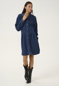 Denim shirt dress, dark blue, with button closure and long sleeves. Features side pockets and tiered design. Worn with black cowboy boots.