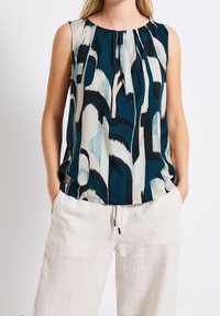 Ärmlös topp i teal, svart och vit med abstrakt mönster och veck, kombinerad med löst sittande vita linnebyxor med snörning och fickor.