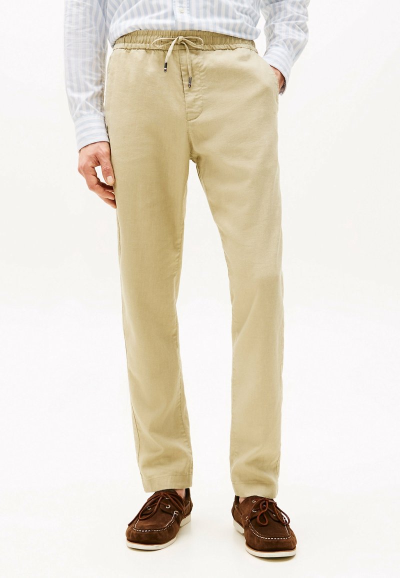 Beige Leinenhose mit elastischem Bund und Kordelzug. Glatte Textur mit lockerer Passform, kombiniert mit braunen Wildlederschuhen im Boot-Stil.
