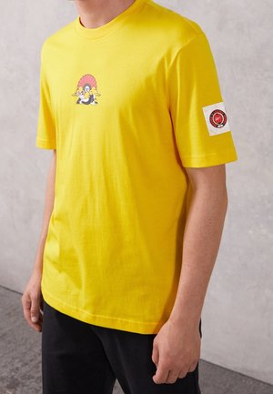 Camiseta amarilla de algodón con un pequeño gráfico de dibujo animado en el pecho, mangas cortas y una etiqueta de logotipo en la manga izquierda.