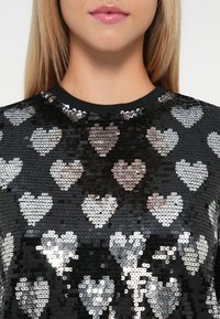 Detailný záber osoby v čiernom top s flitrami, ktorý má lesklé strieborné srdcové vzory a okrúhly výstrih.