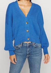 Blå stickad cardigan med djup V-ringning, långa puffärmar och fem stora guldknappar, kort längd, ribbat mönster, tillsammans med jeans.