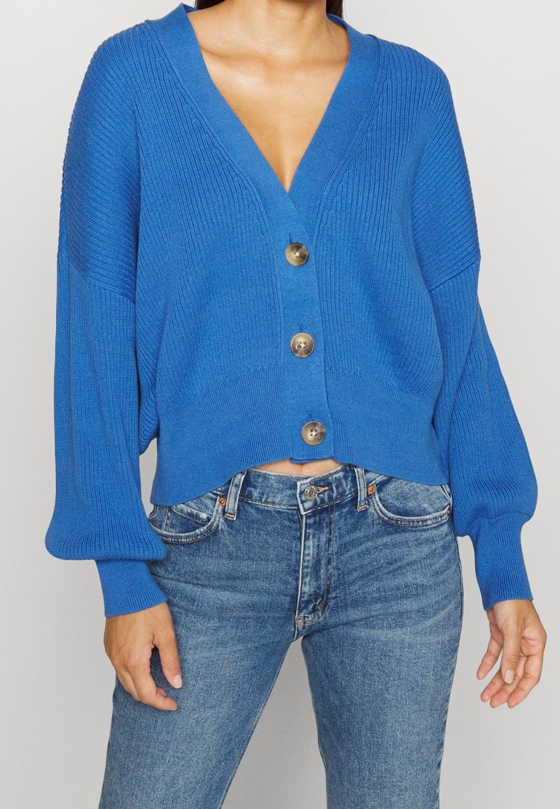 Blå stickad cardigan med djup V-ringning, långa puffärmar och fem stora guldknappar, kort längd, ribbat mönster, tillsammans med jeans.