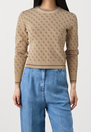 Donna che indossa un maglione beige a maniche lunghe con motivo e pantaloni di jeans a vita alta azzurri chiari su sfondo neutro.