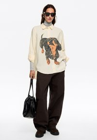 Camisa holgada de color crema claro con estampado de dachshunds en negro y naranja, combinada con pantalones de pierna ancha marrón oscuro y un bolso negro brillante.