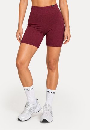 Femme portant un short cycliste bordeaux taille haute avec un motif subtil, des chaussettes blanches avec texte et des baskets argentées, debout sur un sol blanc.