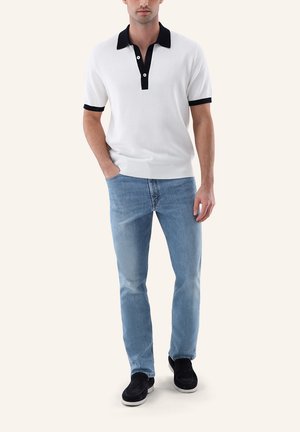 Mann trägt weißes kurzärmeliges Polo mit schwarzem Kragen und Manschetten, hellblaue Jeans und schwarze Slipper, steht mit einer Hand in der Tasche.