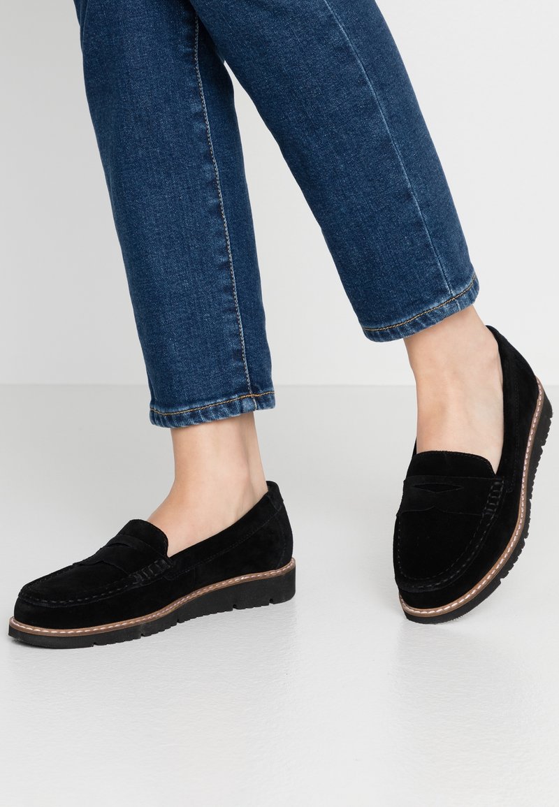 Pieds chaussés de mocassins en daim noir avec semelles marron et noires, associés à un jean court en denim bleu sur fond neutre.