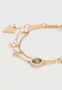 Βραχιόλι από χρυσή αλυσίδα με πολλαπλά νήματα, διακοσμημένο με κρυστάλλινες λεπτομέρειες, ανθικά γούρια και κεντρικό μενταγιόν με λογότυπο, περιτριγυρισμένο από στρας.