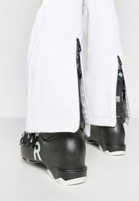 Pantaloni albi de zăpadă cu fermoare laterale, asortați cu bocanci negri de schi care au un logo argintiu, suprafețe texturate și un branț alb.