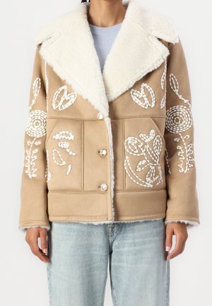 Woman wearing a tan shearling jacket with white embroidered floral patterns, large collar, and silver buttons, paired with light blue jeans.