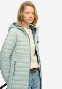 Lyseblå polstret jakke med hætte, der har en fuld lynlås, sidelommer og en tekstureret cremefarvet sweater under. Casual denimjeans.