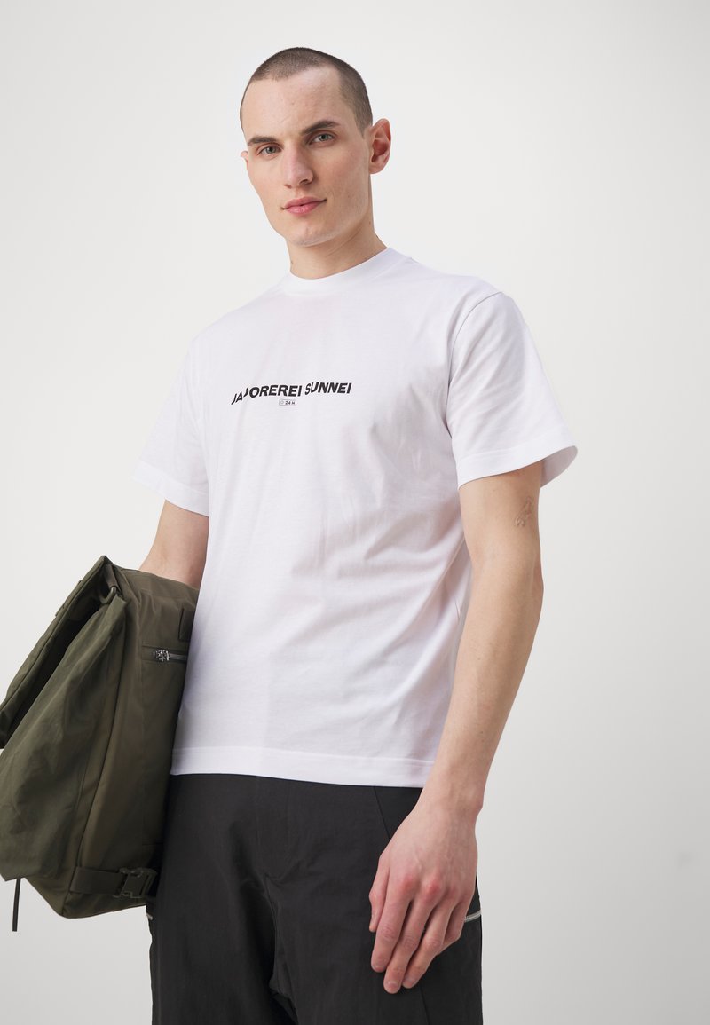 T-shirt bianca a maniche corte con un logo nero al centro. Il modello tiene in mano una giacca verde oliva. Taglio semplice, texture del tessuto liscia.