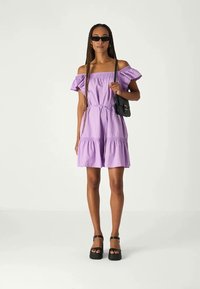 Robe lavande à épaules dénudées avec des manches à volants, taille élastique et jupes superposées. Associée à des sandales noires à plateforme et un petit sac.