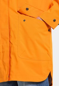 Orange vattentålig jacka med en slät textur, en stor framficka och tryckknappar på ärmarna. Har sidoveck för att underlätta rörelse.