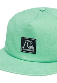 Green cotton cap with a flat brim, featuring a black square patch with white wave graphic and text "QUIKSILVER." Soft, textured fabric.