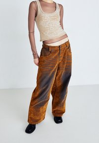 En tanfärgad ärm- och ärmlös tröja över en brun mesh långärmad tröja, parat med löst sittande, orange-bruna corduroybyxor med ljusare vertikala ränder och fem knappar.