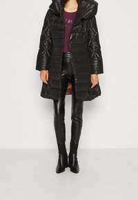 Giacca imbottita nera con cappuccio, trama trapuntata e lunghezza a metà coscia. Indossata sopra pantaloni aderenti in pelle nera e stivali alla caviglia con tacco.